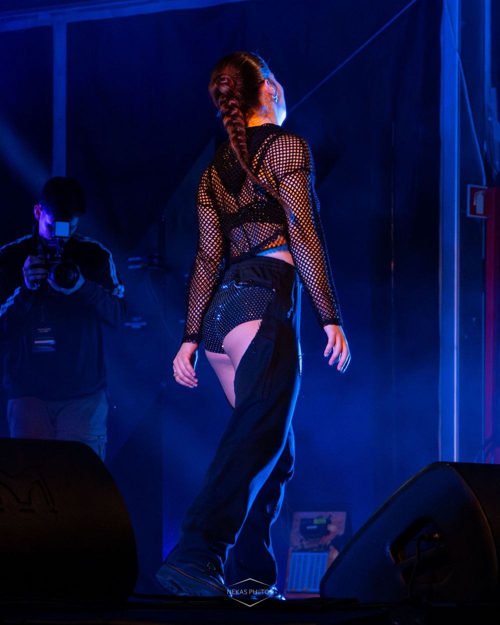 Female performer in black fishnet top and sequined pants on a blue-lit stage with a photographer in the background.