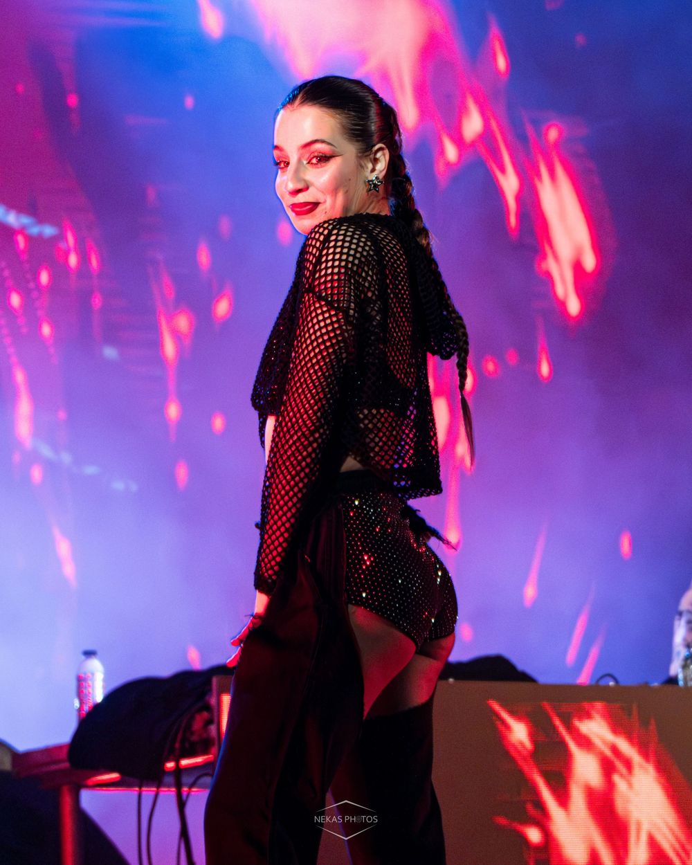 Female performer in black mesh top poses on stage with vibrant red and purple light display behind her.