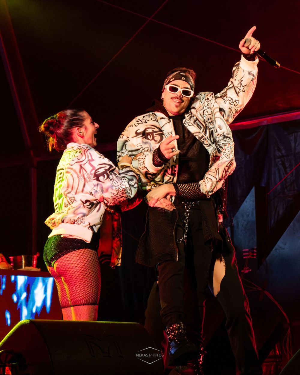 Two performers in graphic print jackets on stage, one pointing up, under red lights at a live music event.