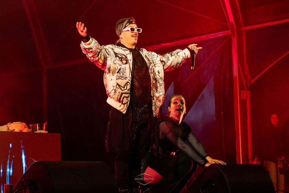 Energetic performer in graffiti jacket spreads arms wide on red-lit stage with dancer crouching dramatically behind.