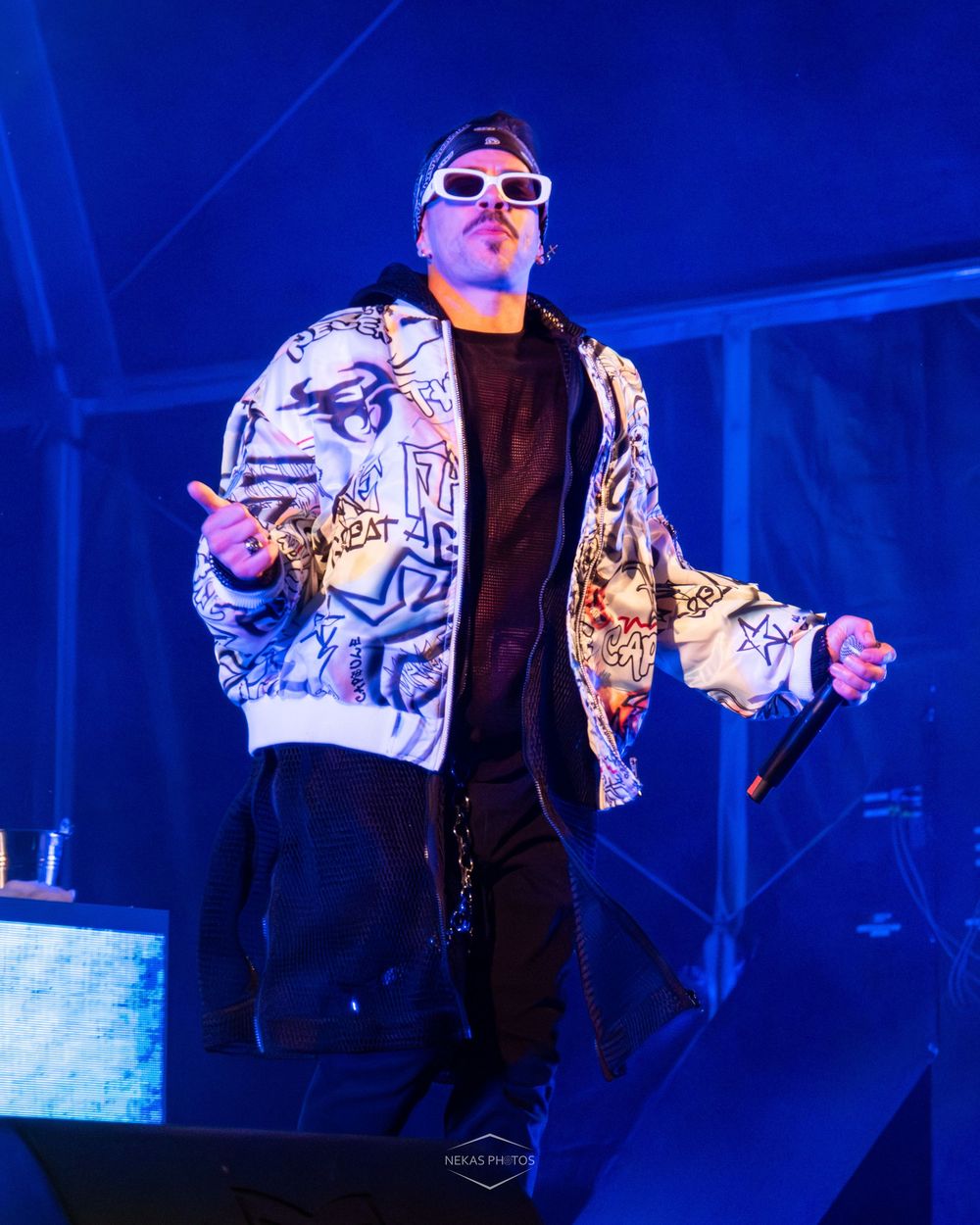 A performer in a graffiti-print bomber jacket and white sunglasses holds a microphone on a blue-lit stage.