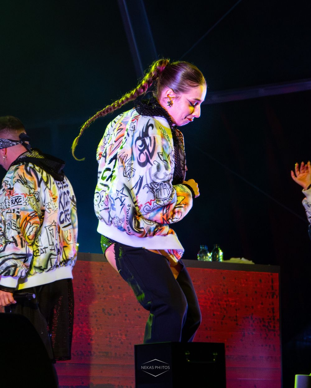 Performer in graffiti print jacket with long braid dancing on stage under colorful lights.