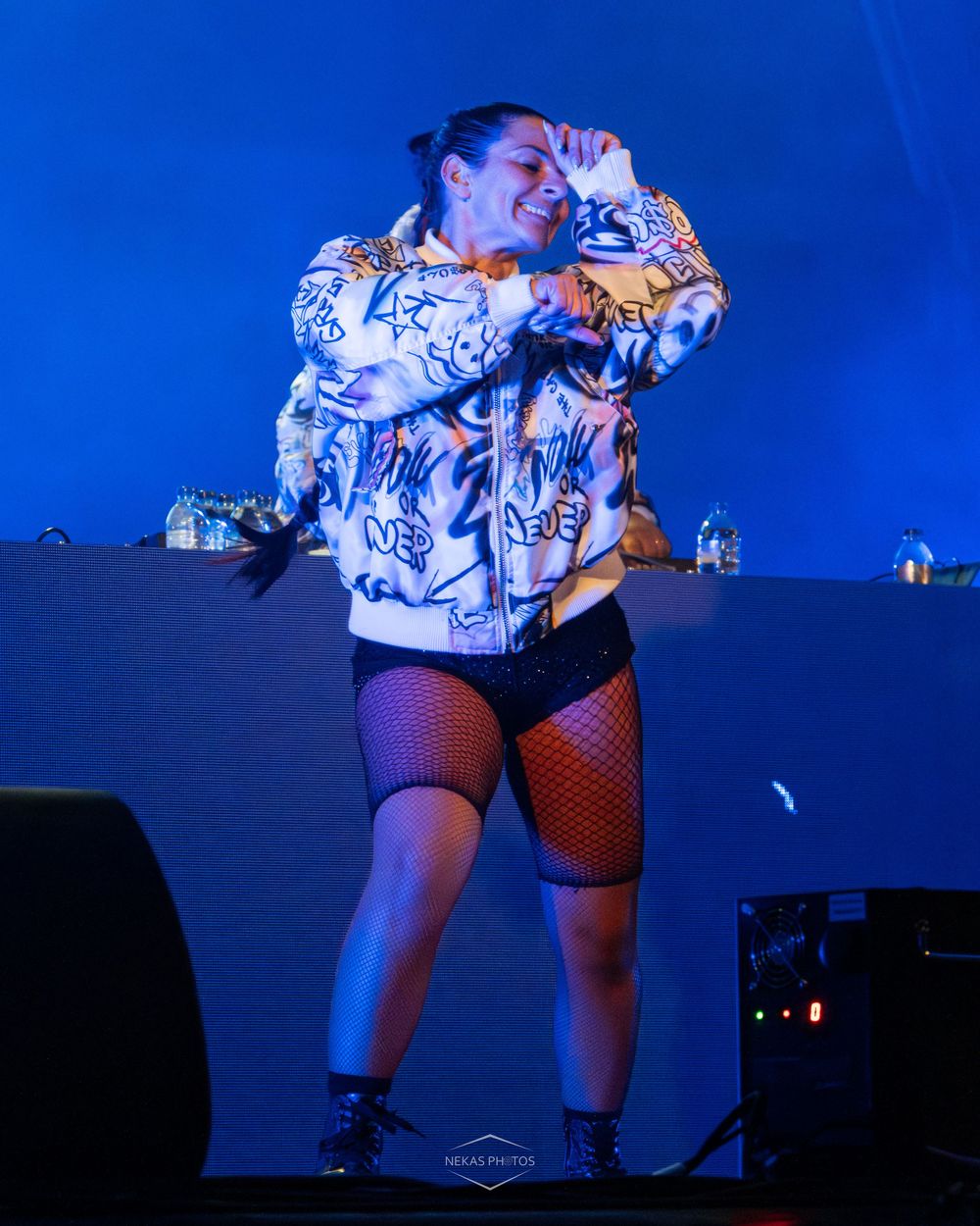 Female DJ performing on stage wearing graffiti jacket and fishnet stockings under blue stage lighting.