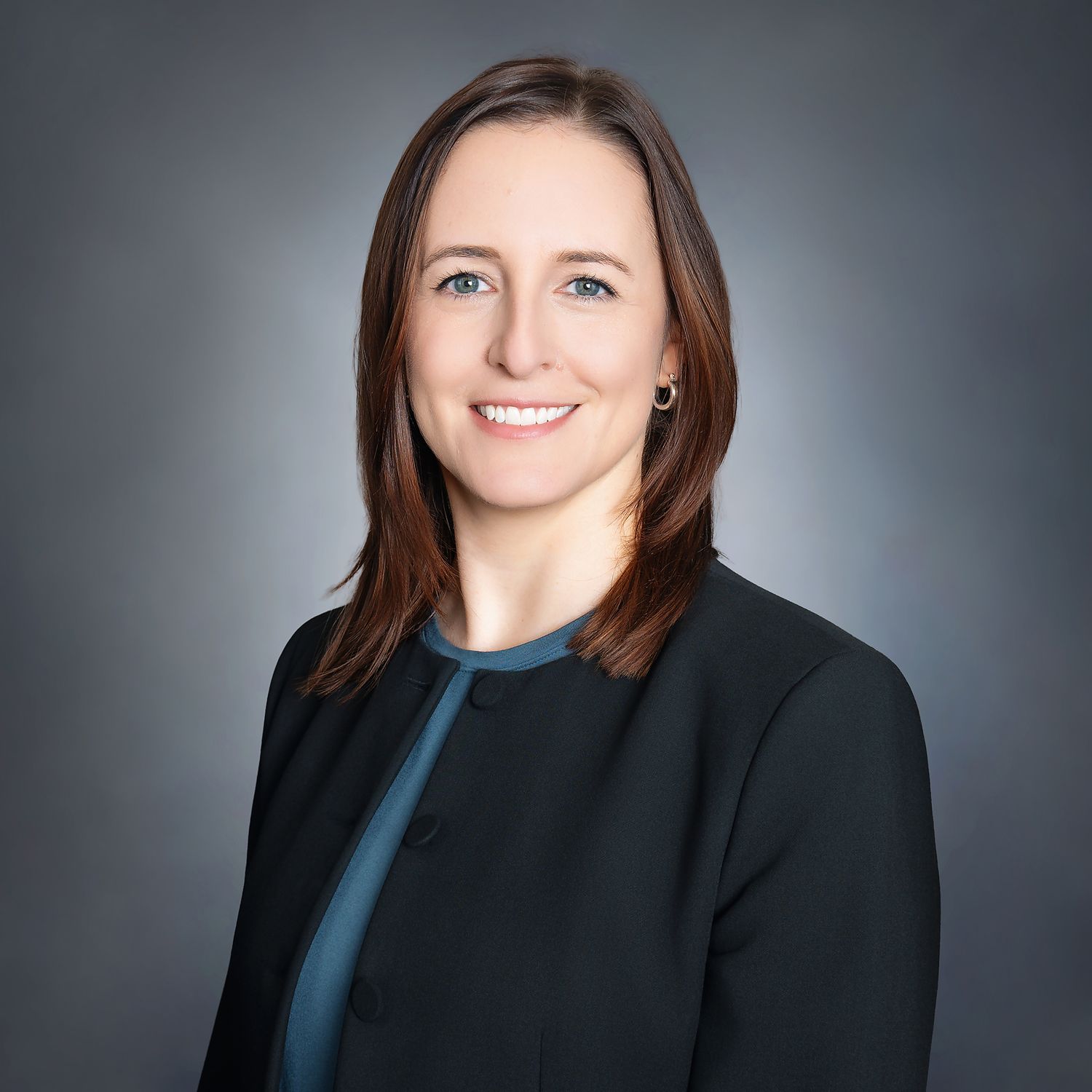 Professional headshot of a woman with brown hair wearing a dark blazer and teal top against a gray background.