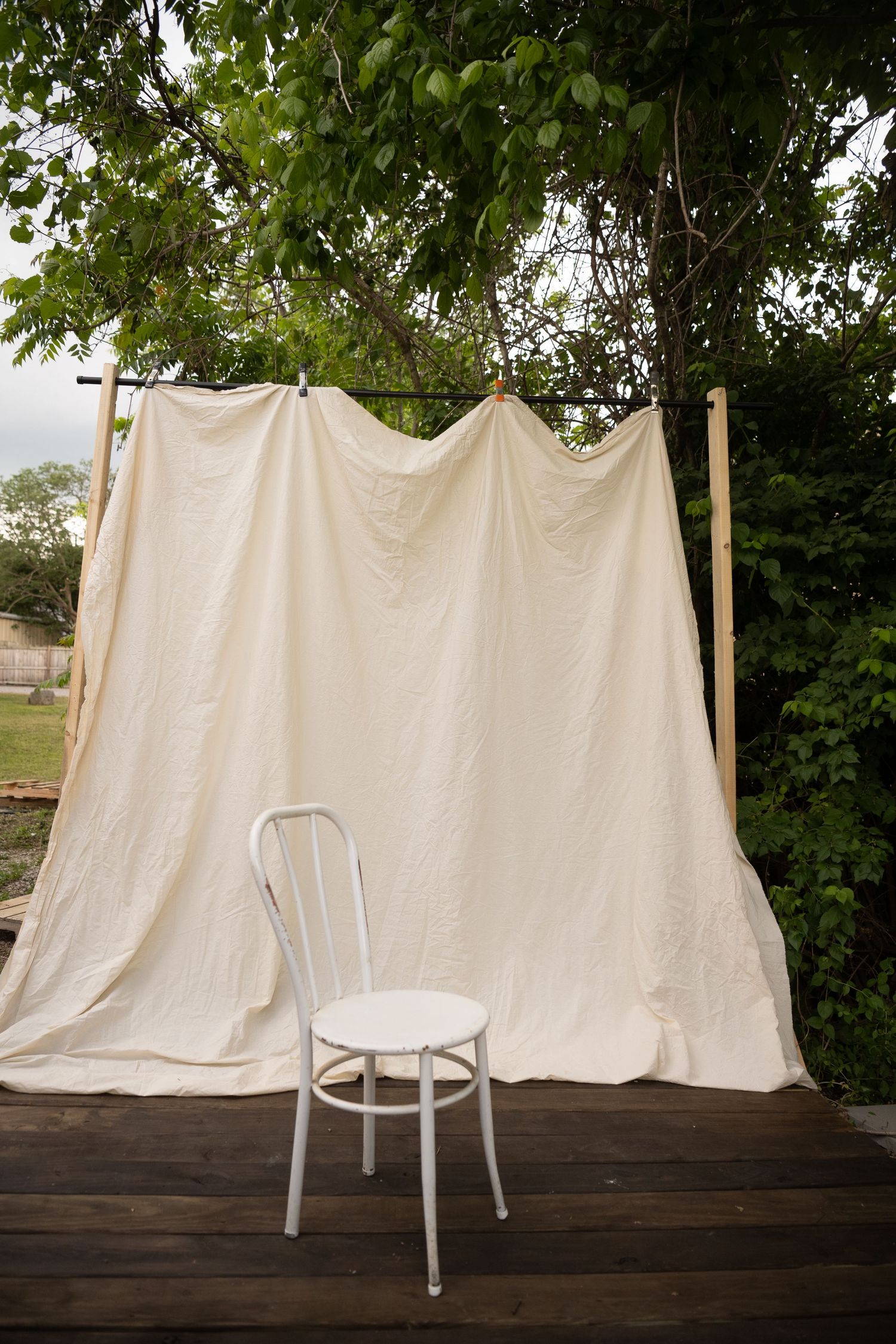 White chair on wooden deck with cream fabric backdrop hung between wooden posts outdoors.