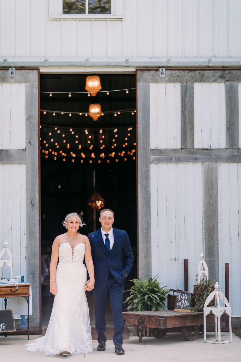 Bride and groom holding hands outside a rustic white barn adorned with string lights at their wedding reception.