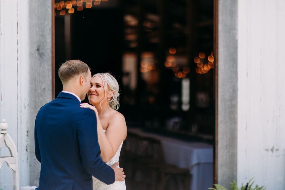 Bride and groom share a tender moment outside a rustic barn venue, with the groom whispering to his smiling bride.