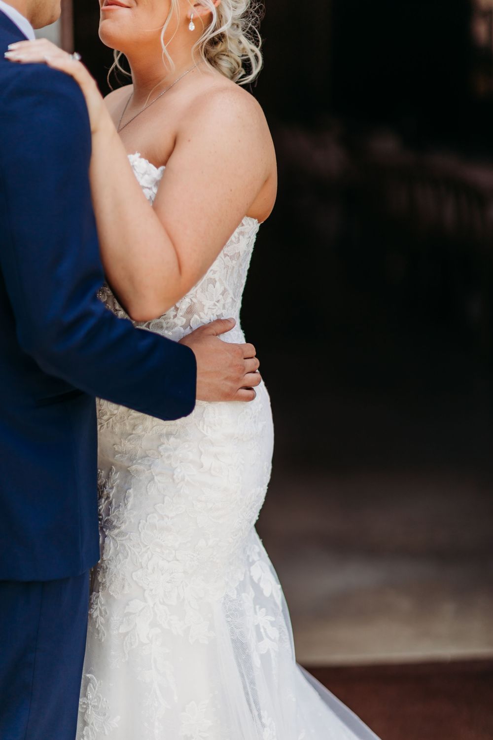Bride in a lace mermaid wedding gown embraced by groom in navy suit during outdoor wedding portrait.