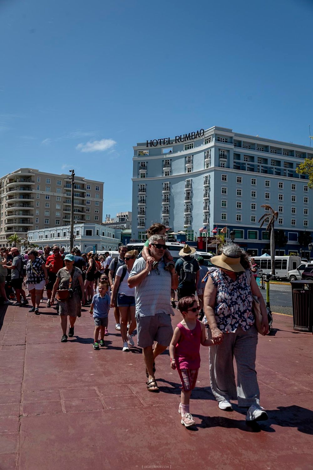 Crowds of tourists walk along a waterfront promenade with Hotel Rumbo visible in the background under a clear blue sky.