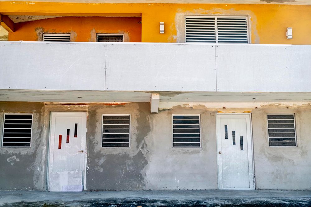 Weathered concrete building exterior with white doors, barred windows, and yellow upper facade.