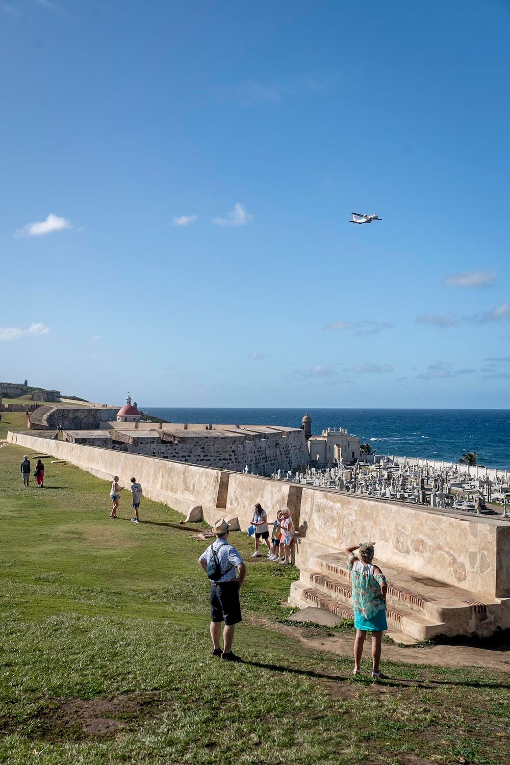 Tourists explore the historic walls of El Morro fort in San Juan, Puerto Rico, with a plane flying overhead and ocean views.