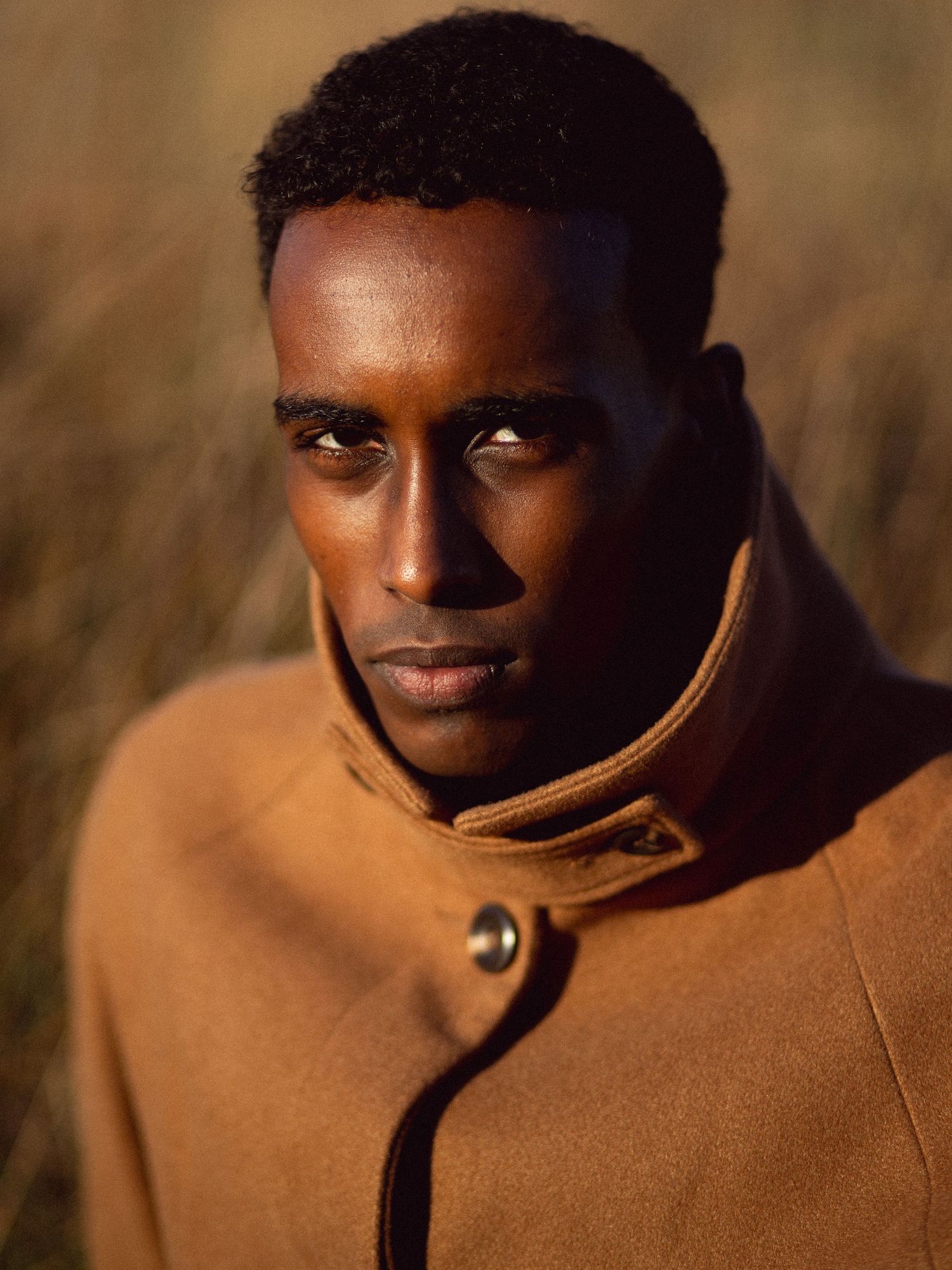 A young Black man in a camel wool coat poses outdoors against a blurred golden grass background.
