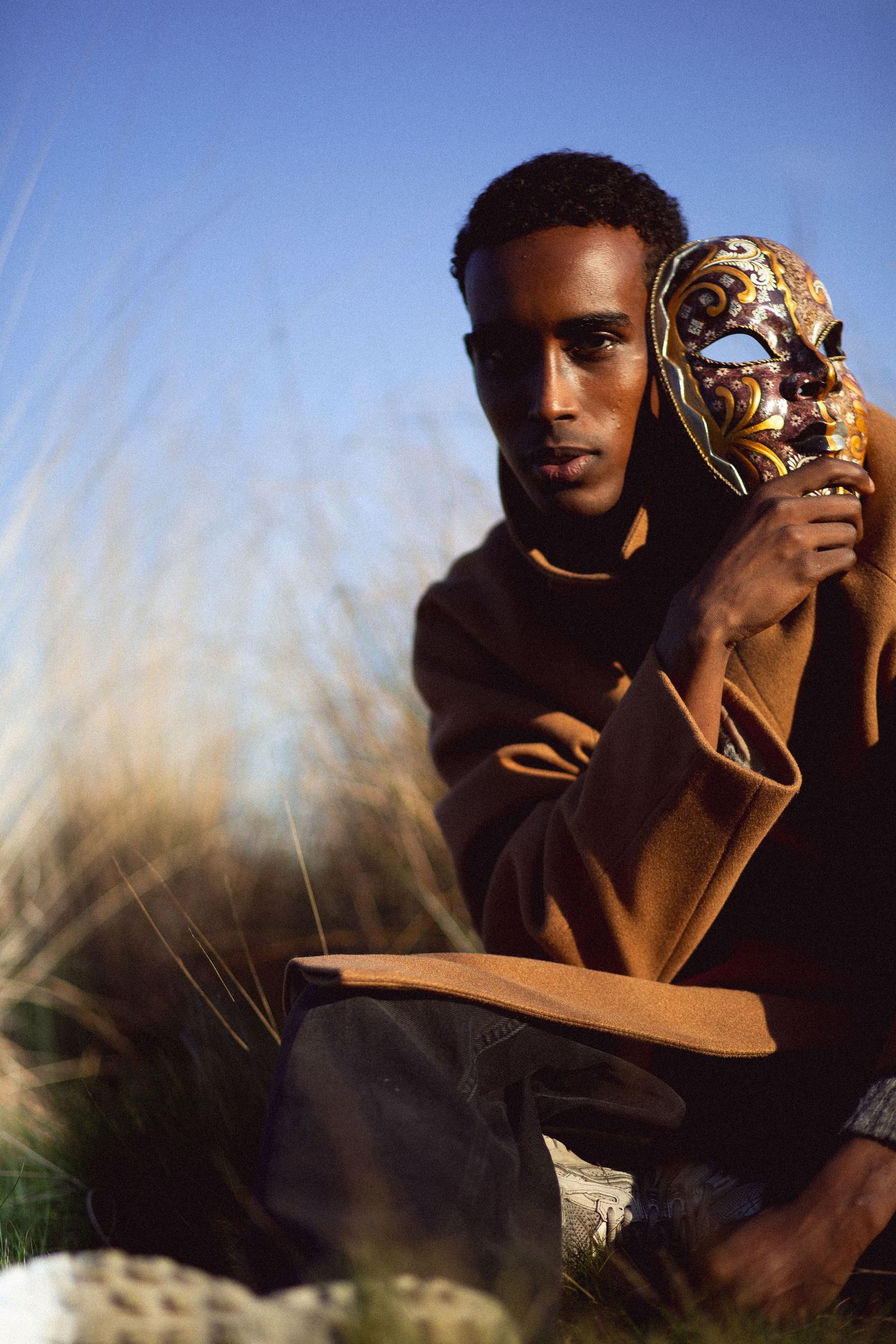 Young Black man in brown coat holds ornate Venetian mask outdoors among tall grass under clear blue sky.