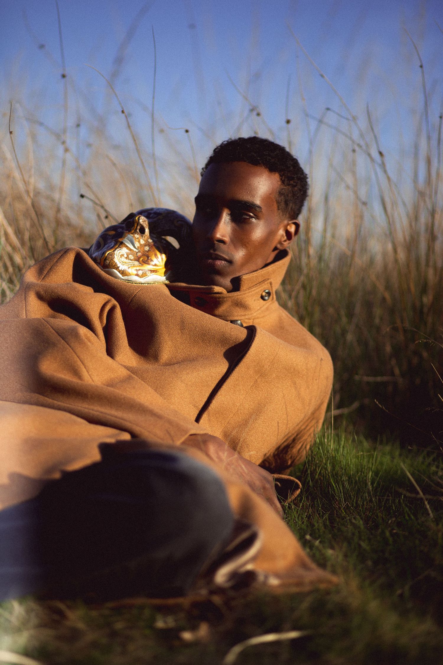 A young Black man wrapped in a brown blanket sits in tall grass outdoors, holding a gold floral accessory.