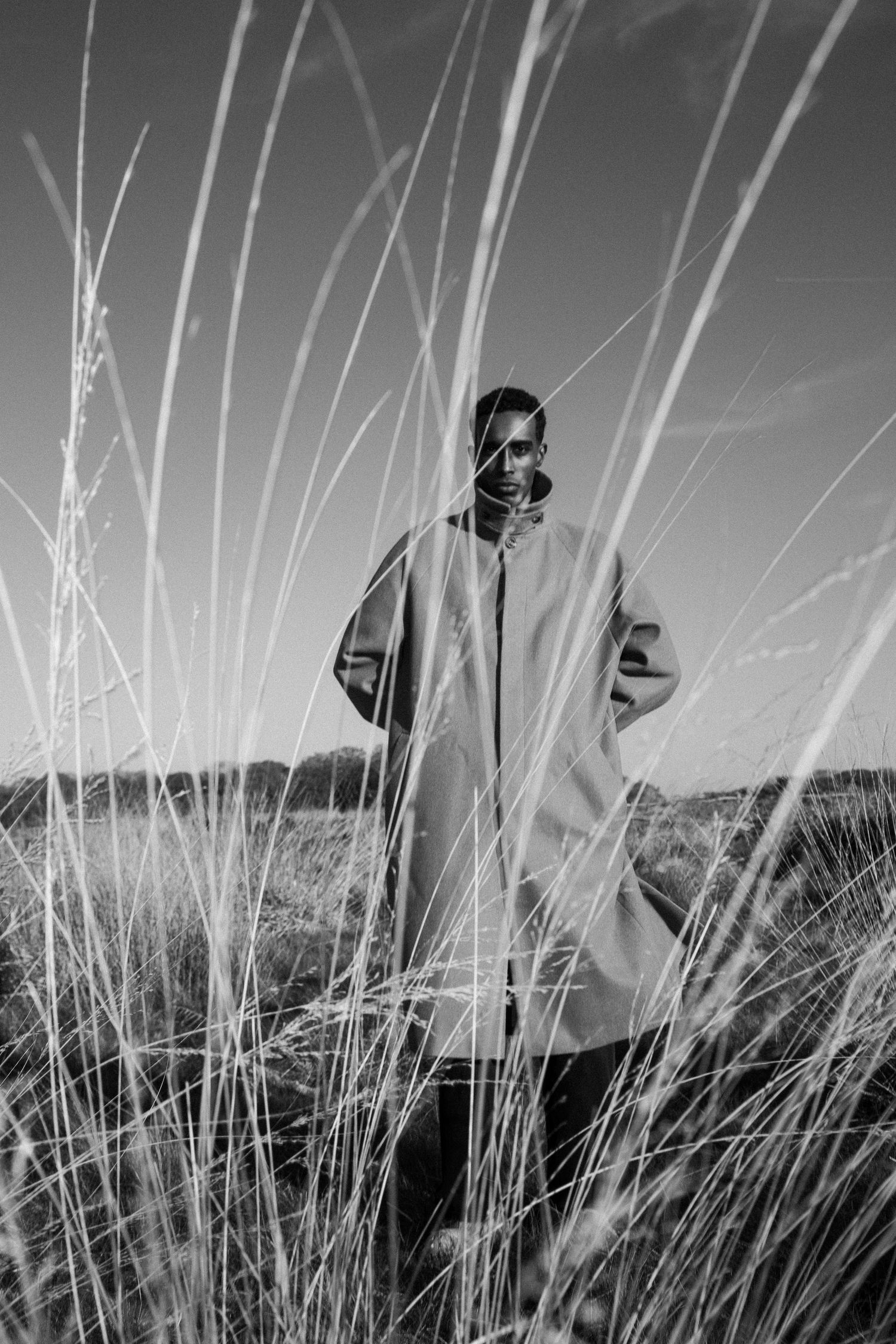 Black and white photo of a person in an oversized coat standing among tall grass in an open field.
