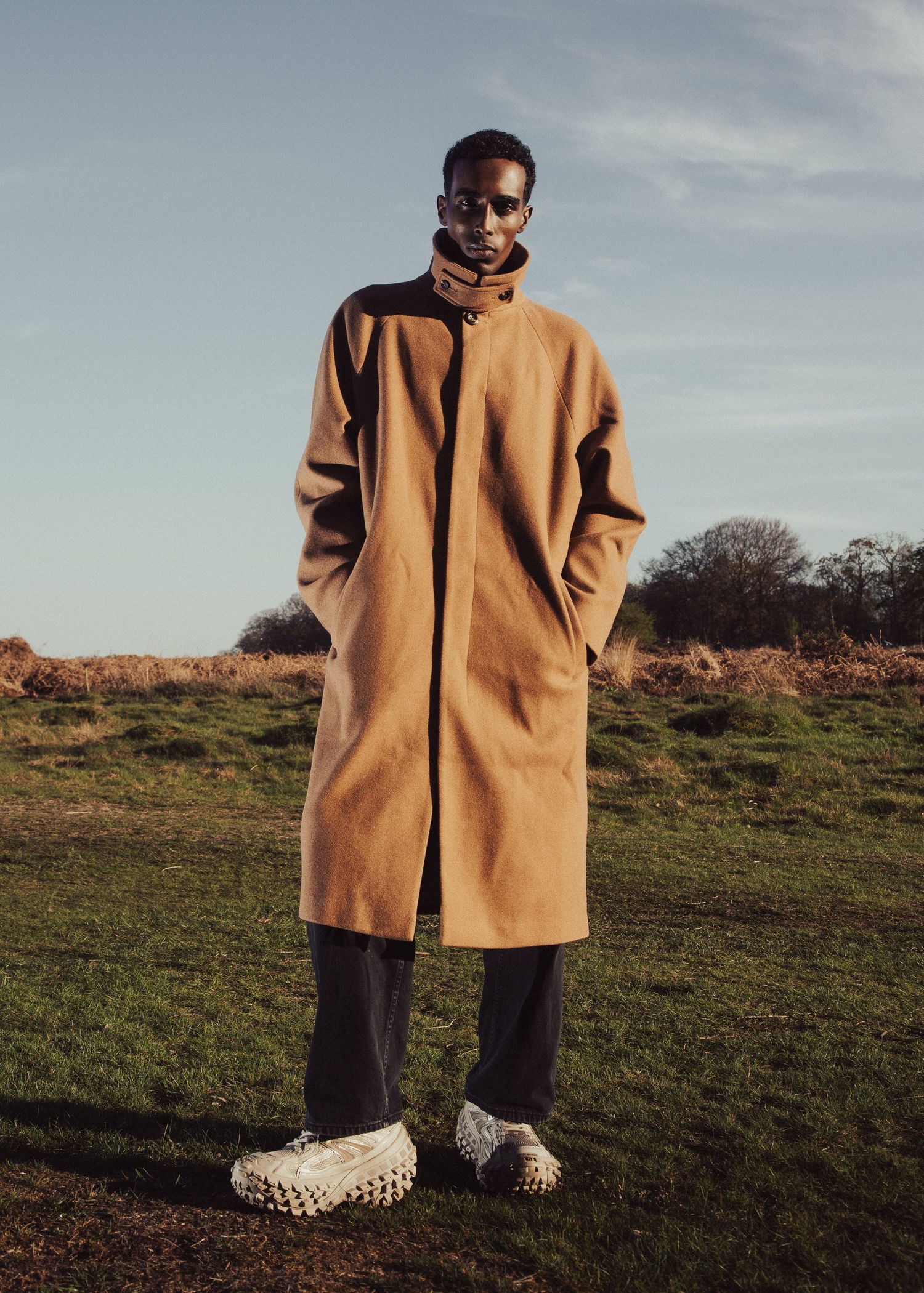 Man wearing a camel longline coat with dark trousers and textured white shoes, standing in a green field.
