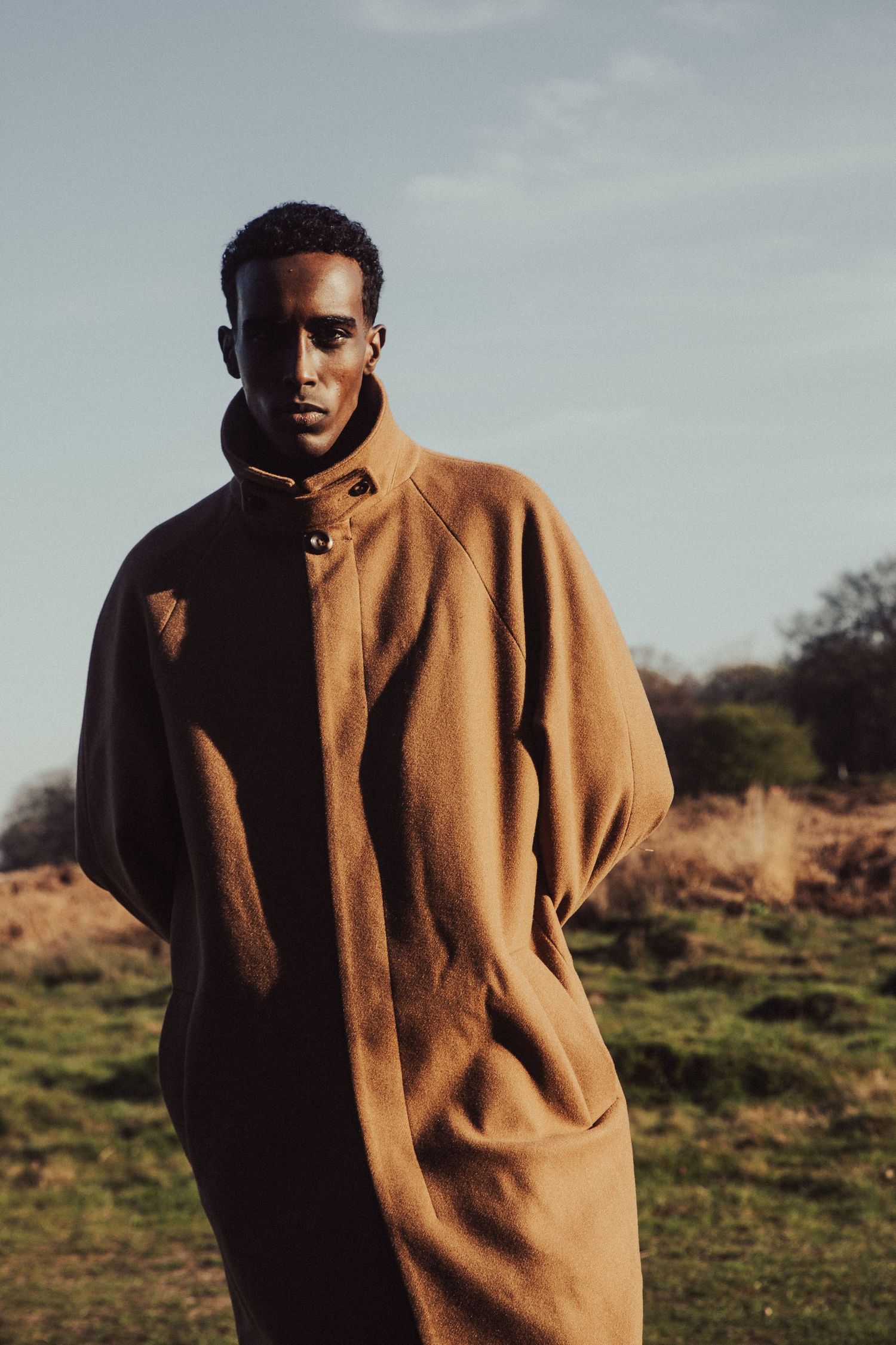 Man in camel oversized wool coat standing in a field with blue sky background.