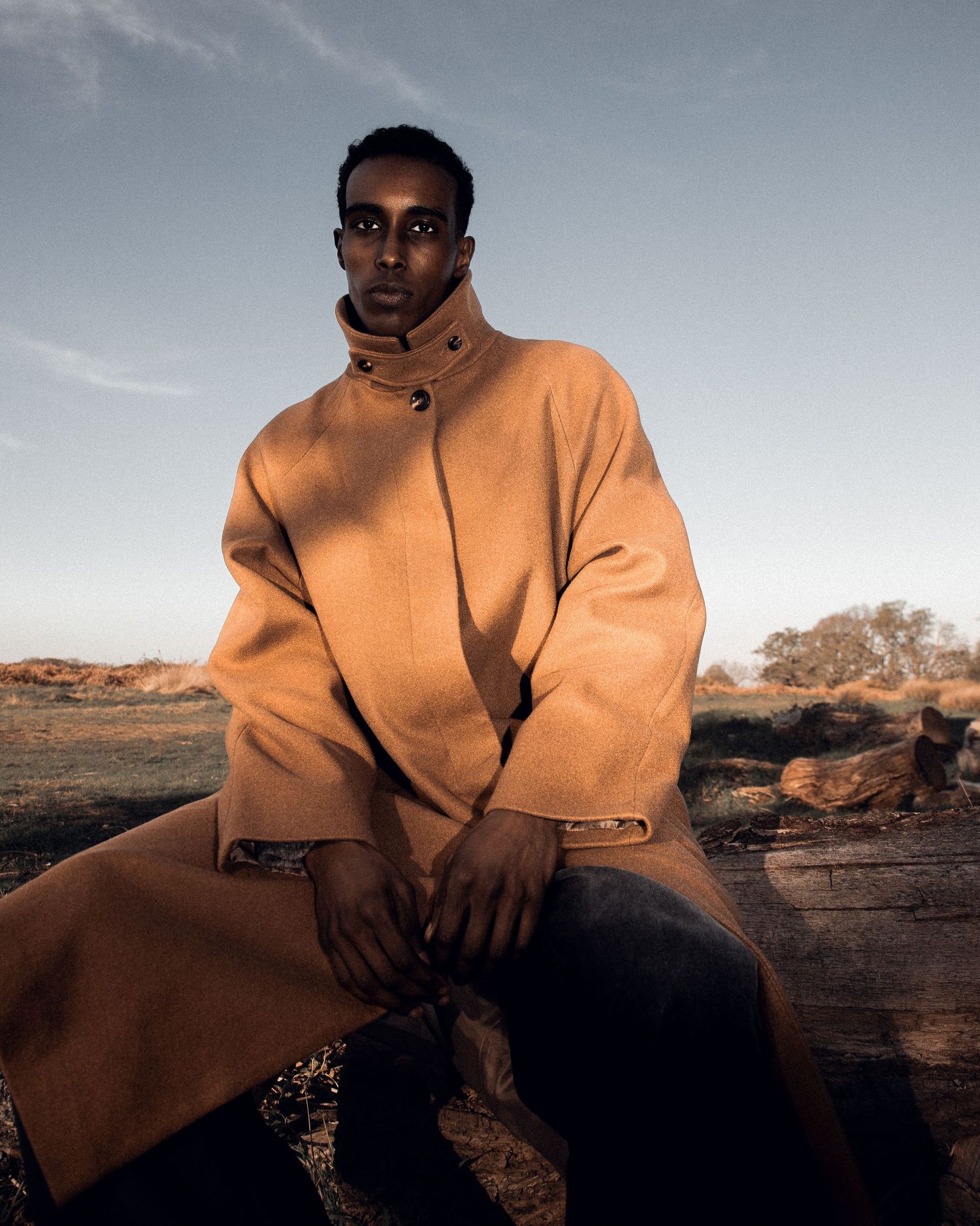 Man in camel oversized coat sitting on rocks in a vast outdoor landscape during golden hour.