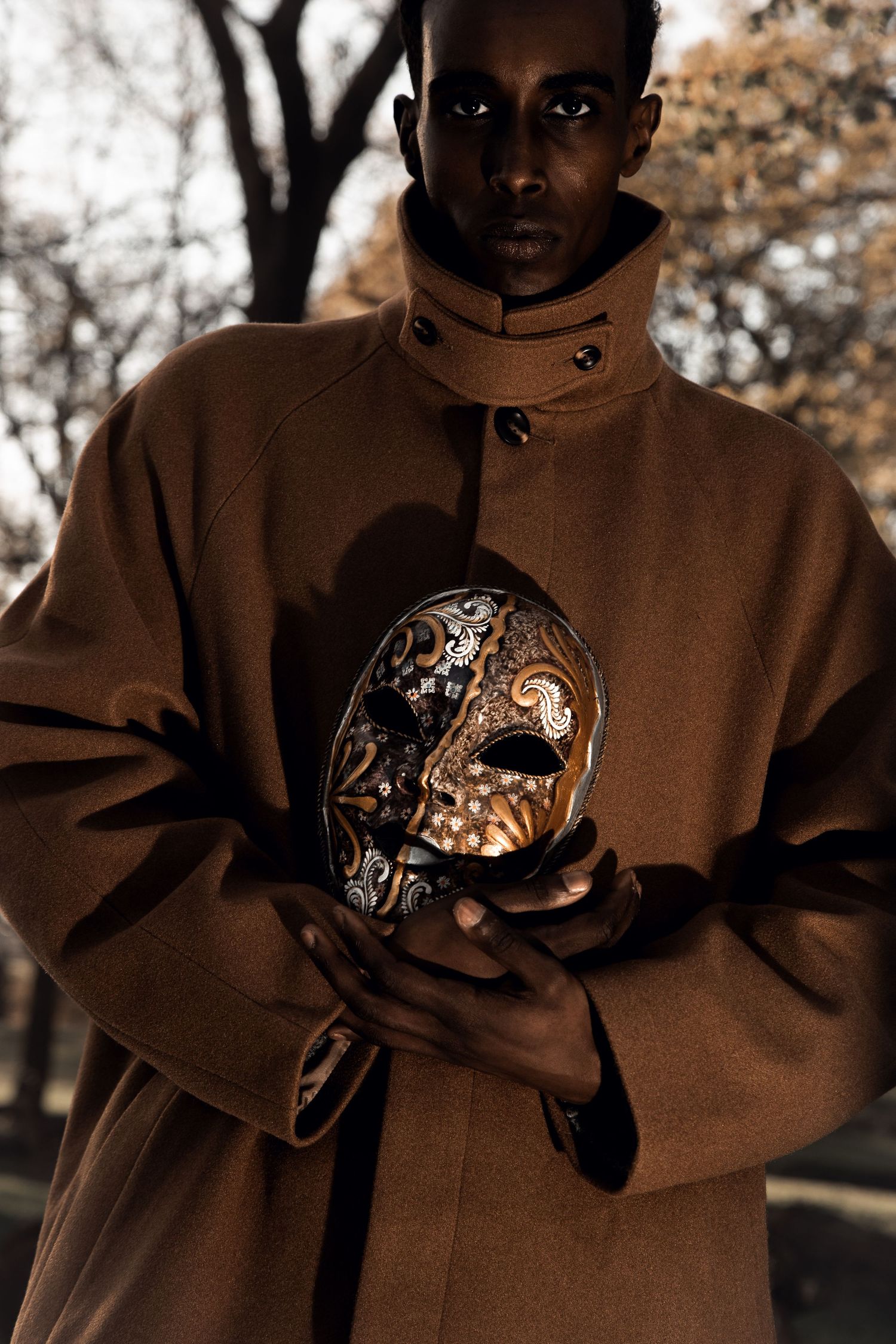 A young Black man in a brown coat holds an ornate Venetian masquerade mask outdoors in autumn.