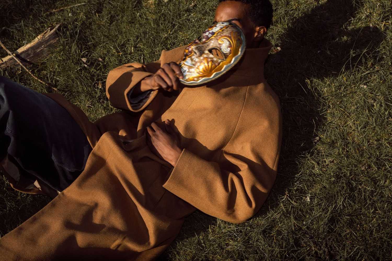 Person in brown coat lying on grass holding a golden mask in warm sunlight.