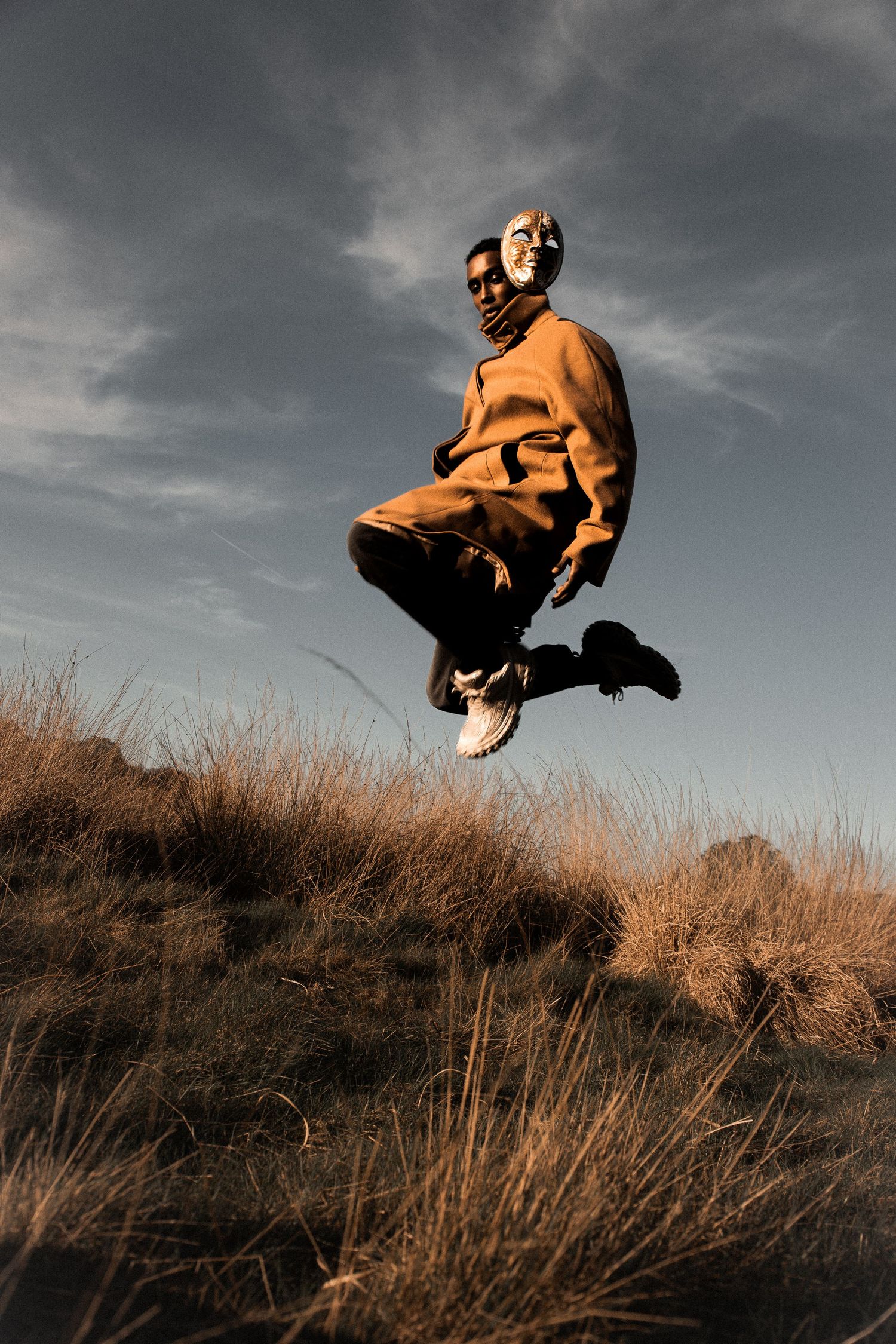 Man in orange hoodie jumping mid-air over dry grassland against dramatic cloudy sky.
