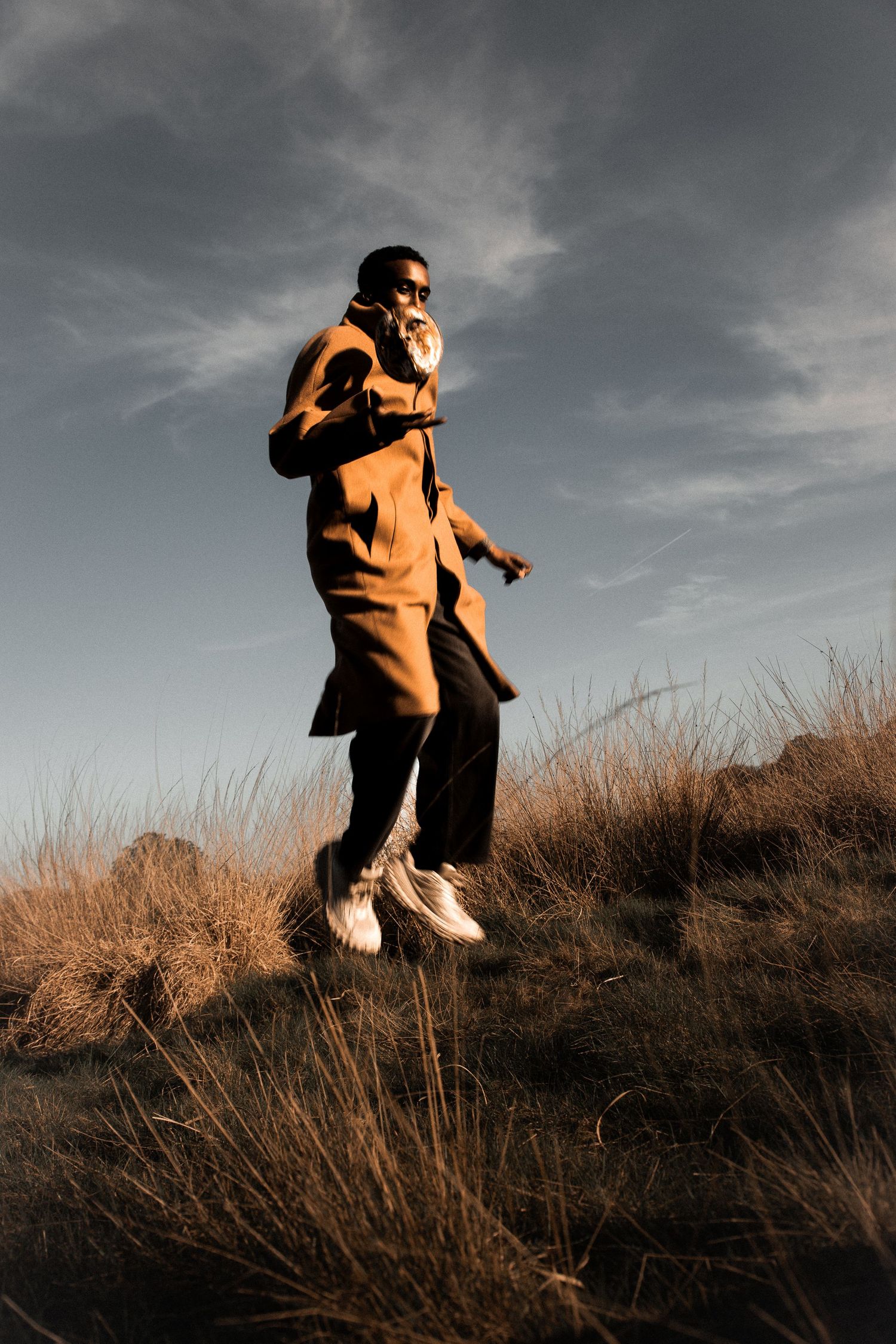 Man in orange coat and white sneakers jumping over dry grass field under dramatic cloudy sky at golden hour.