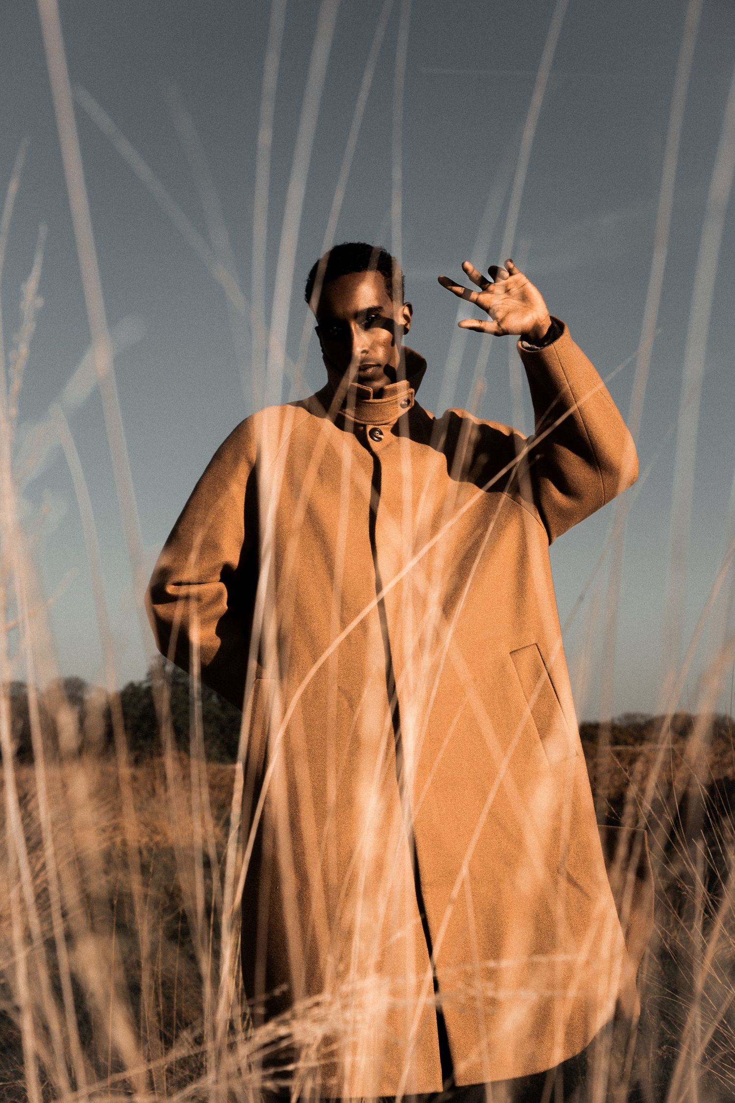 Man in oversized tan coat posing through tall dry grass in golden outdoor light.