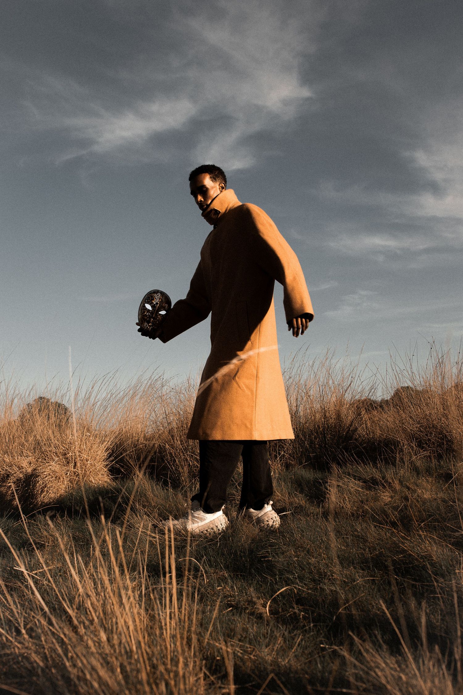 Man in orange apron and black sleeveless shirt holds a dark object in a golden grass field under a cloudy sky.