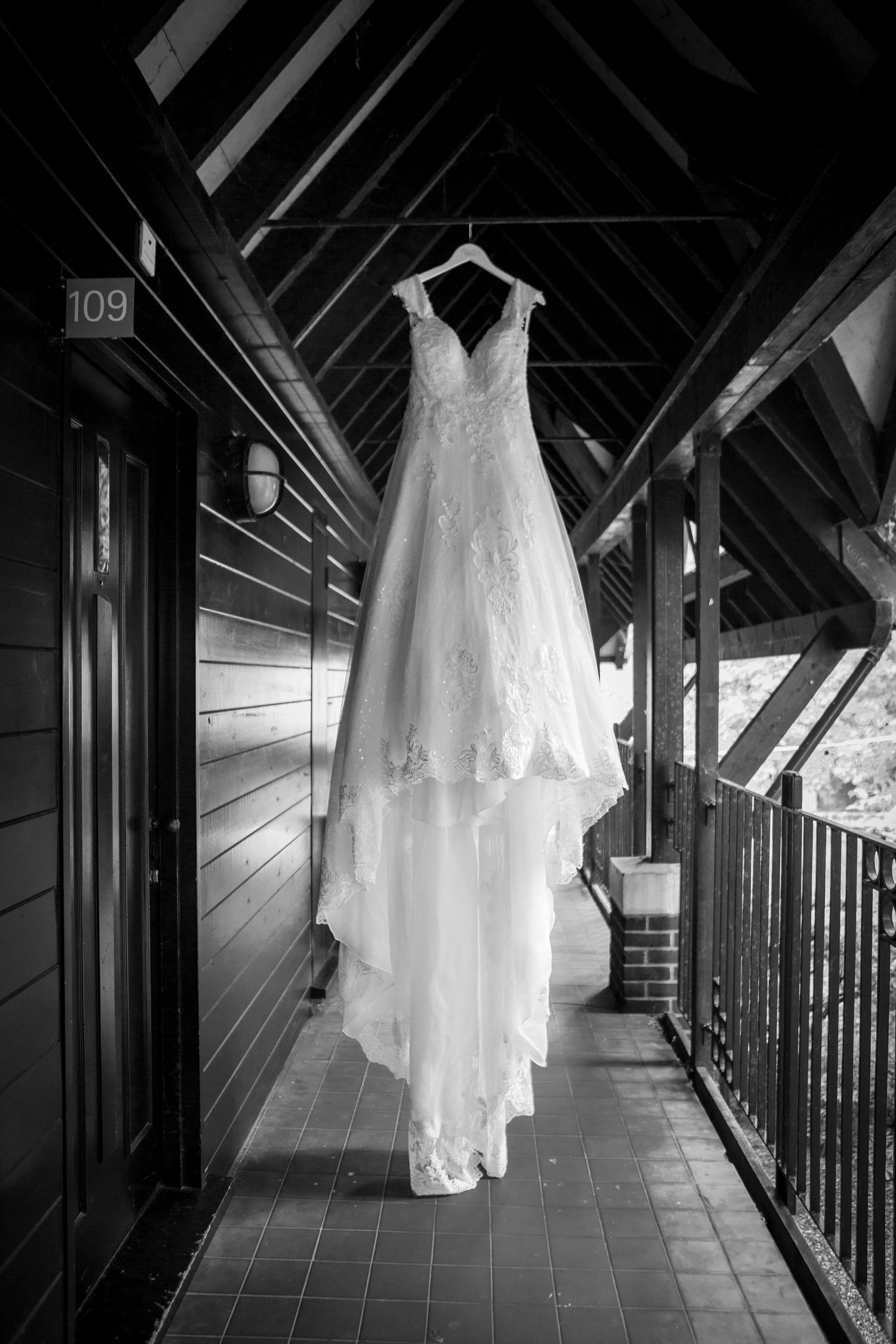Black and white photo of lace wedding dress hanging on outdoor wooden balcony corridor.
