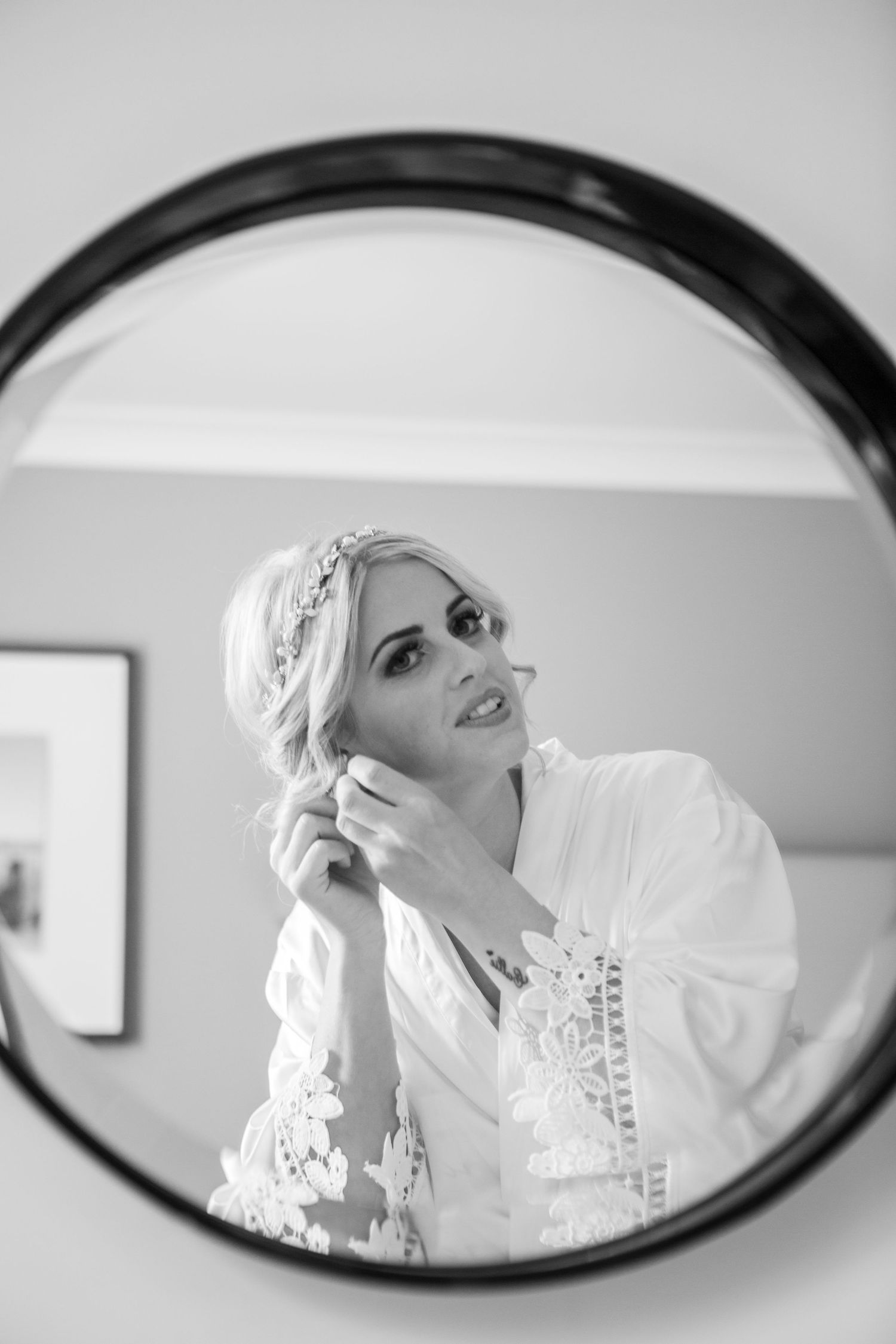 Black and white mirror reflection of blonde bride putting on earrings in white robe.