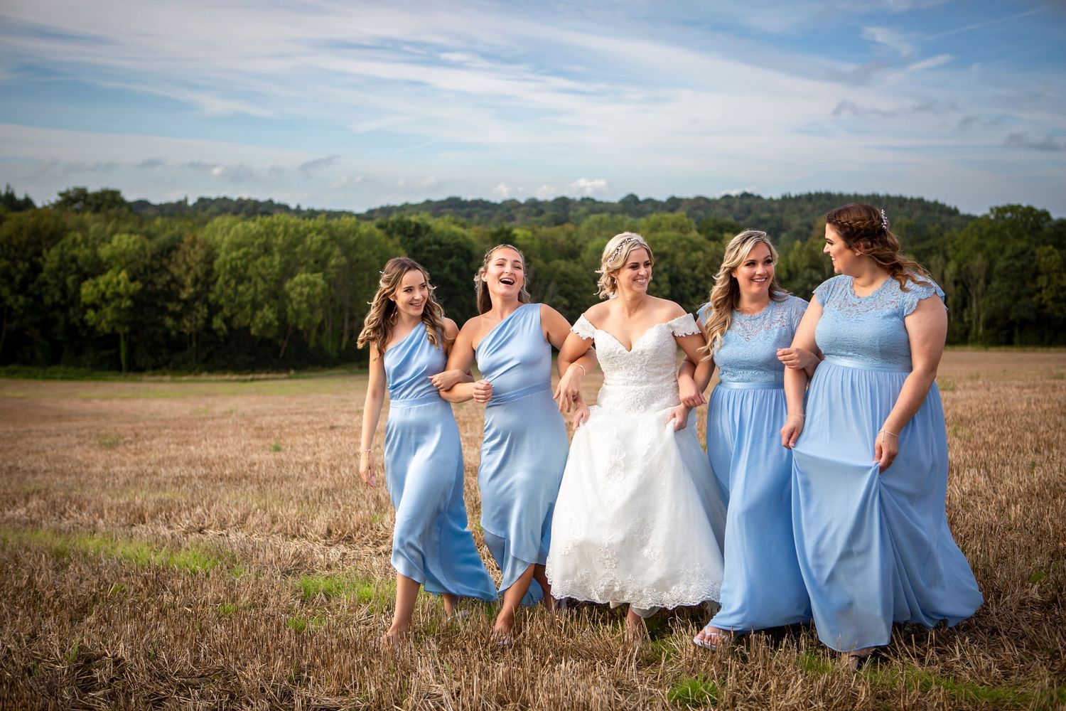 Bride and four bridesmaids in blue dresses laughing while walking through golden harvest field.