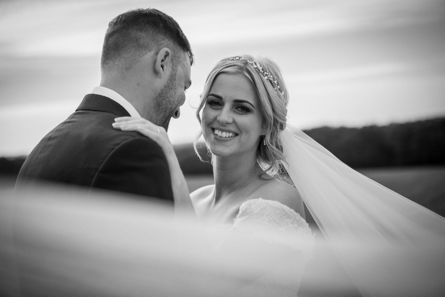 Black and white romantic portrait of smiling bride in veil gazing at groom outdoors.