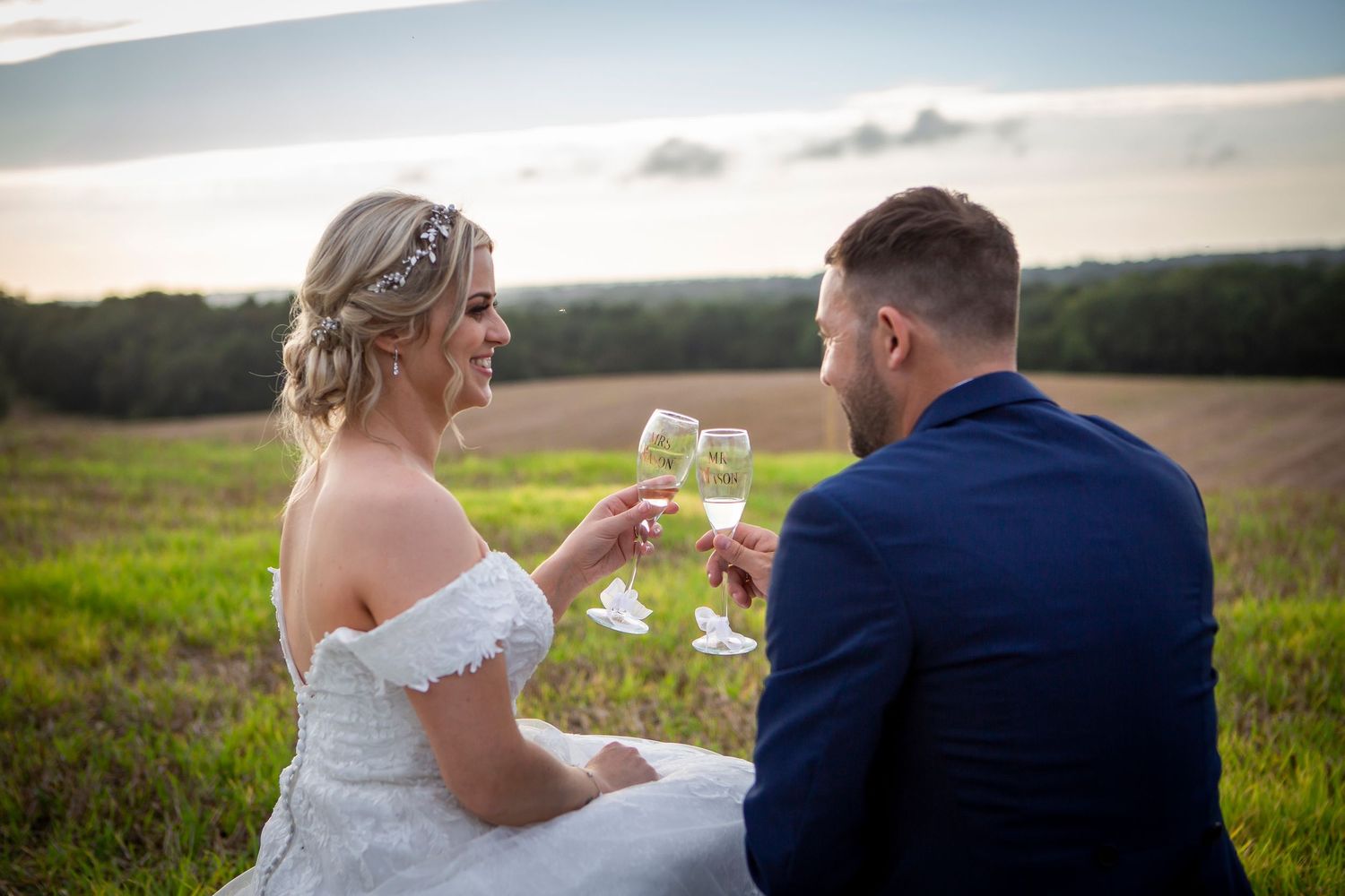 Bride and groom toast with champagne glasses while sitting in a scenic countryside field at sunset.