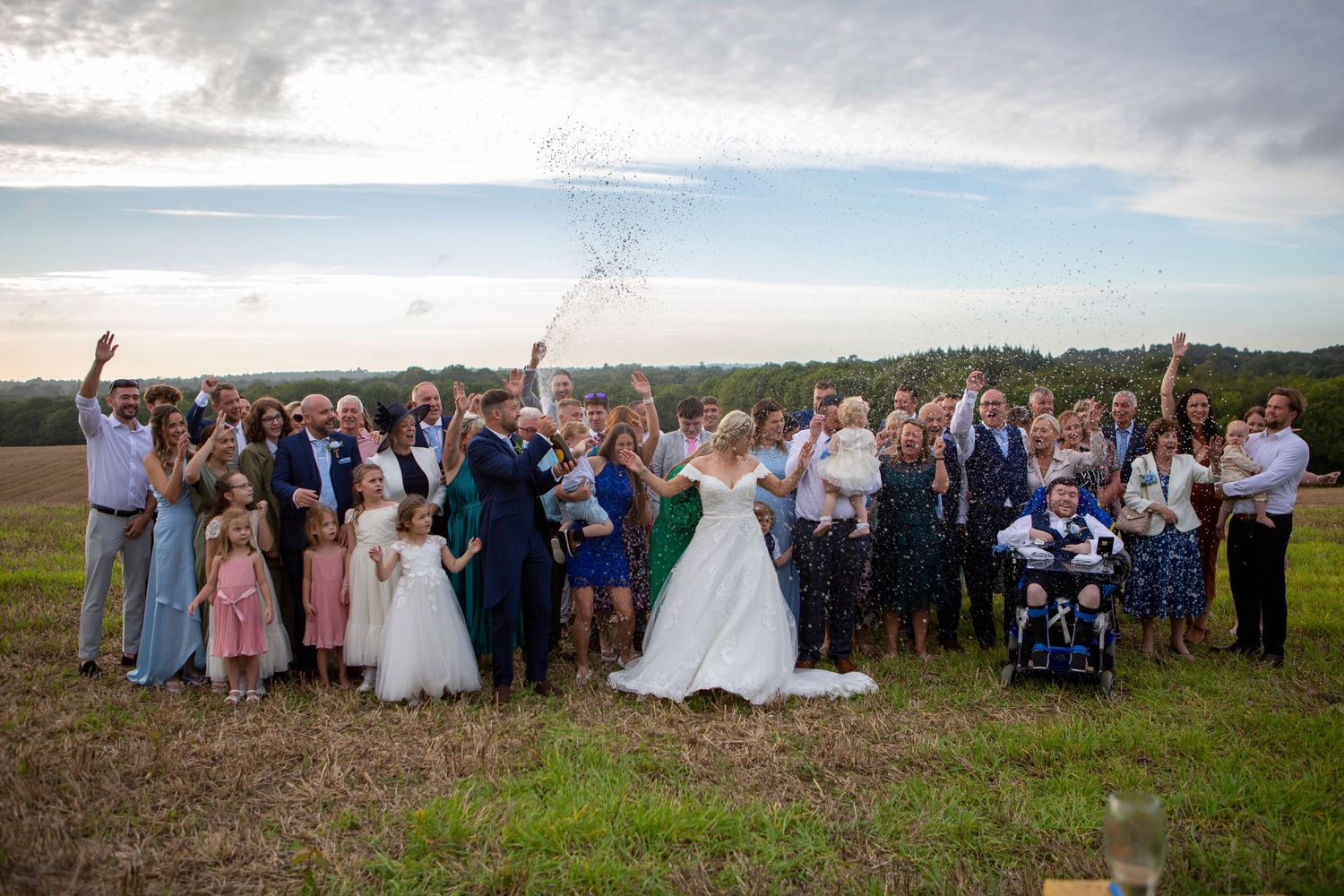Large wedding family group photo celebrating outdoors in countryside field at sunset.