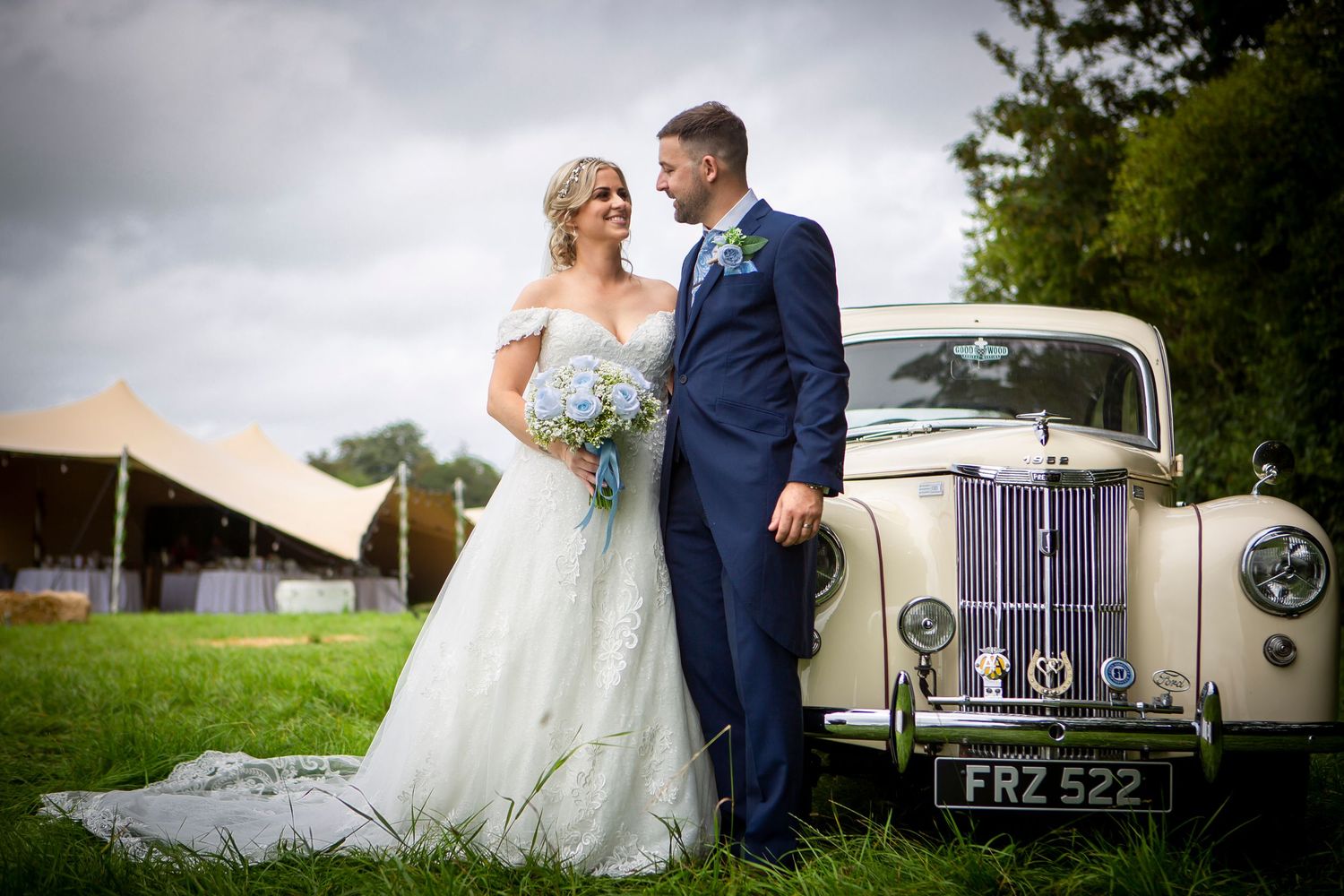 Bride and groom pose beside a vintage ivory wedding car with a tipi venue in the background.