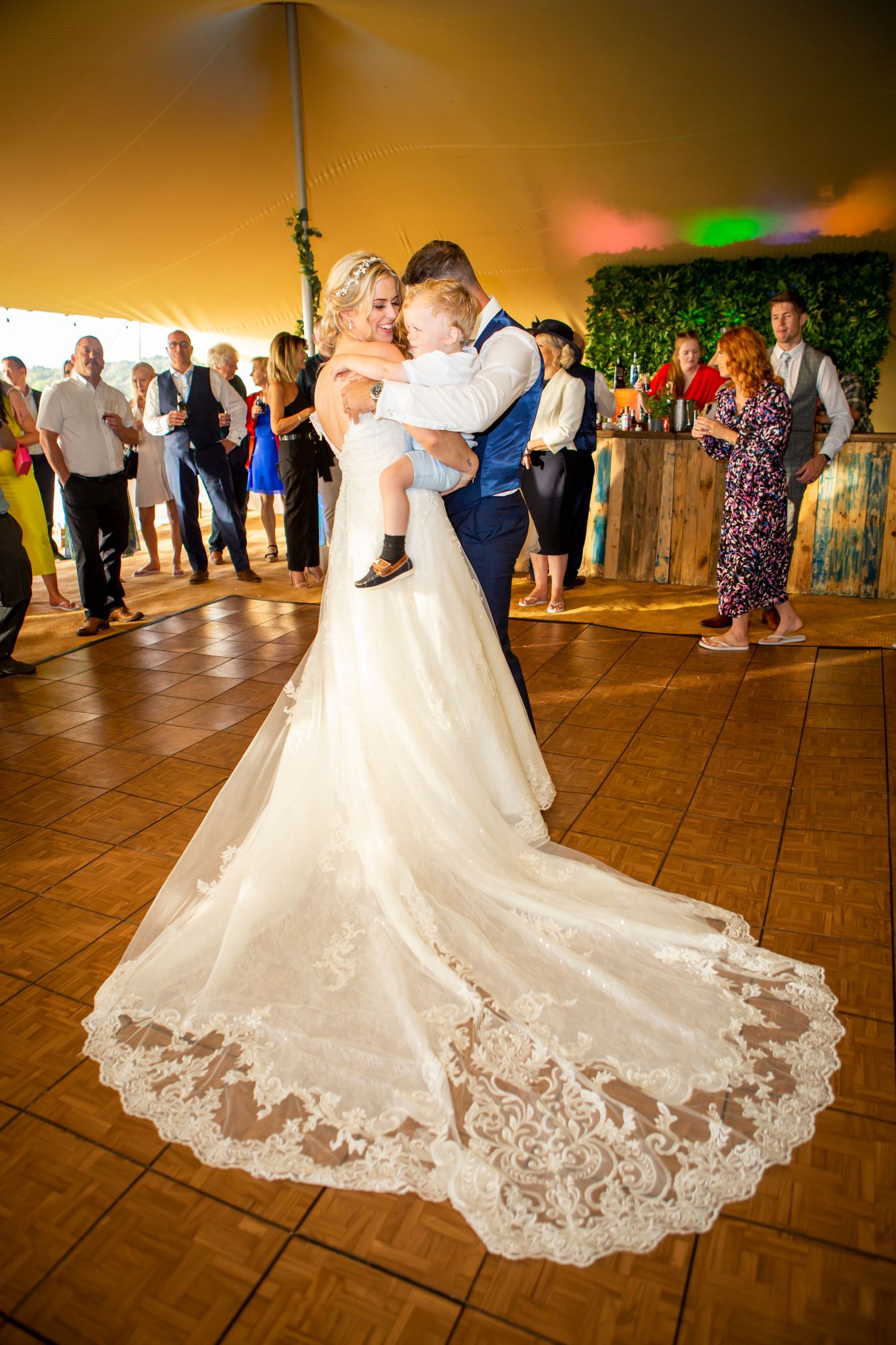Bride and groom share first dance holding toddler with lace train spread on dance floor.