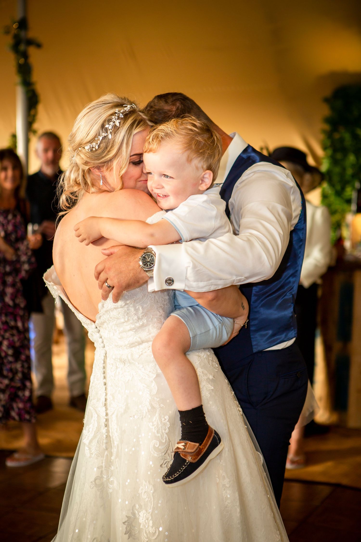 Bride and groom share tender first dance moment while holding young child at wedding reception.