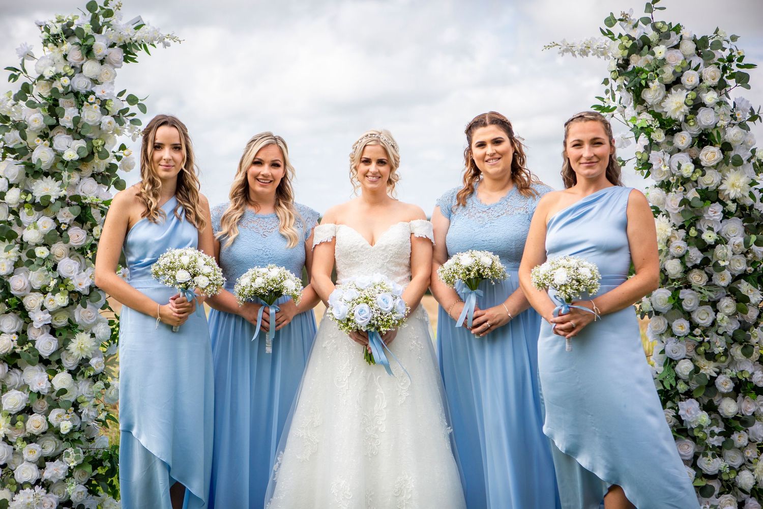 Bride with three bridesmaids in blue dresses holding bouquets in front of white floral arch.