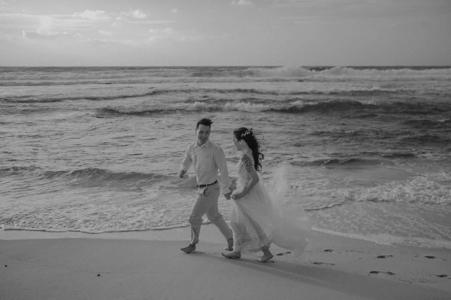 Couple sharing a romantic kiss on a sandy beach in Hawaii, bride holding white bouquet, ocean waves and green cliffs behind.