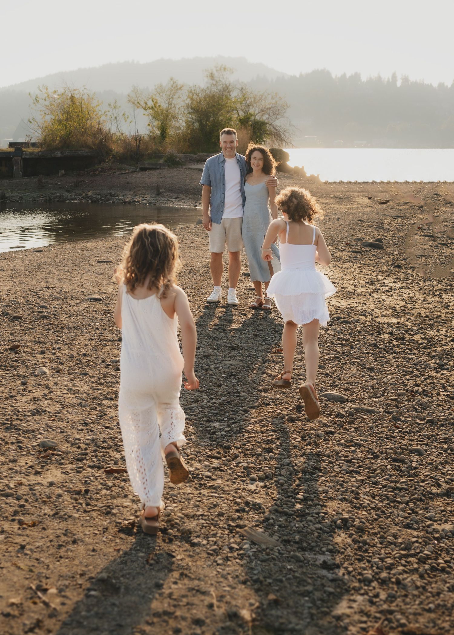Two children in white dresses run toward their parents on a sandy beach at golden hour.