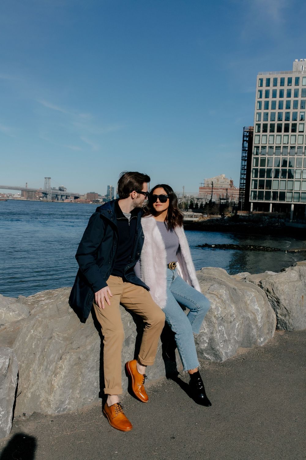 Couple sitting on rocks by the waterfront in NYC, wearing stylish fall outfits with a bridge and modern building backdrop.
