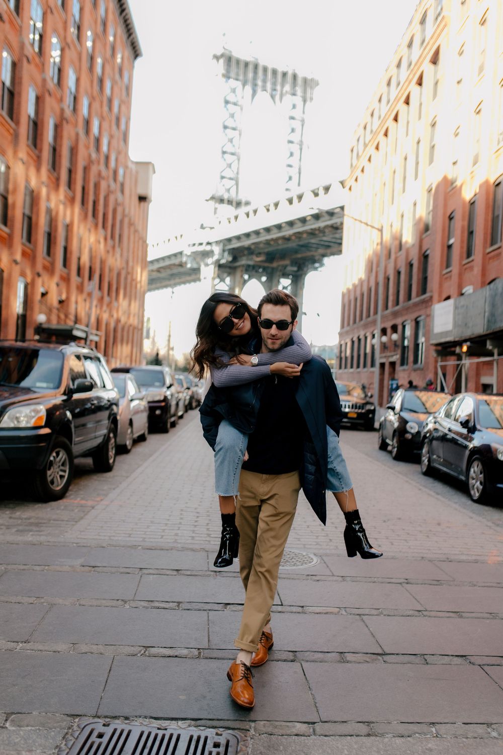 Couple enjoying a piggyback ride on a cobblestone street in DUMBO, Brooklyn, with the Manhattan Bridge in the background.