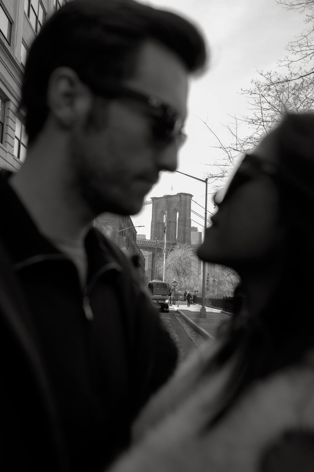 Black and white photo of a couple in sunglasses sharing a romantic moment with the Brooklyn Bridge in the background.