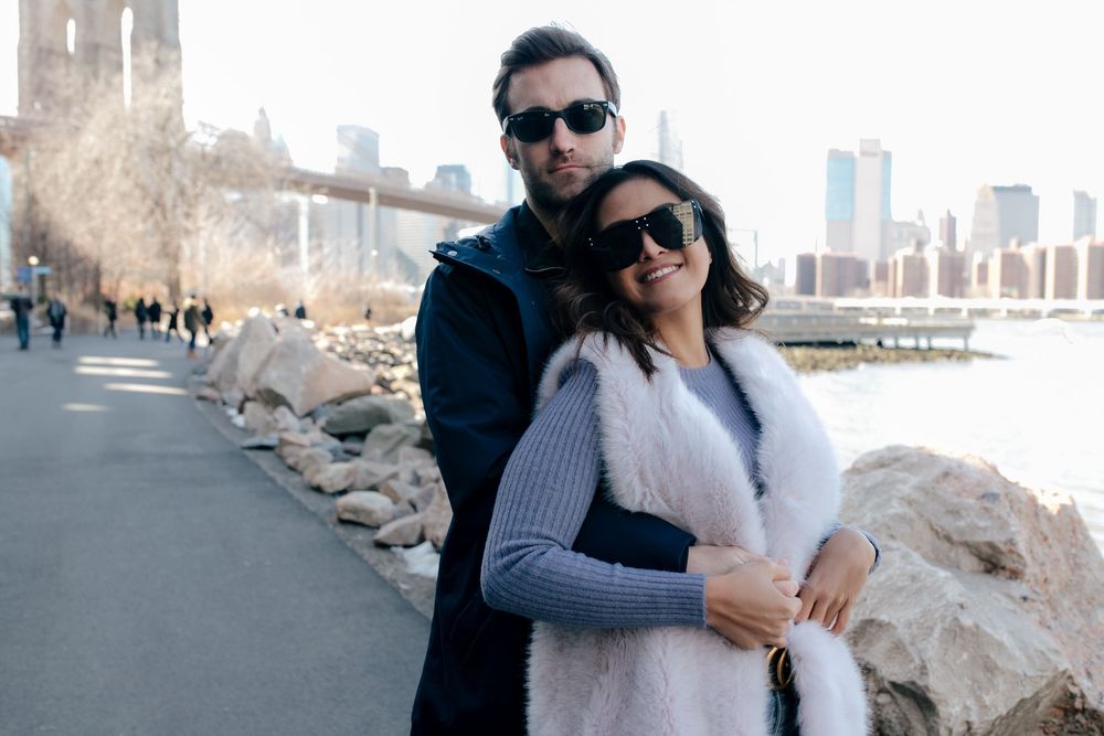 Couple in sunglasses embracing near Brooklyn Bridge waterfront with NYC skyline in winter.