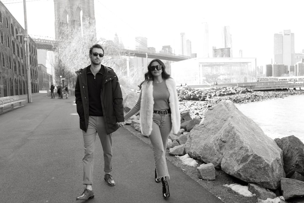 Couple holding hands walking along a waterfront path with the Brooklyn Bridge in the background.
