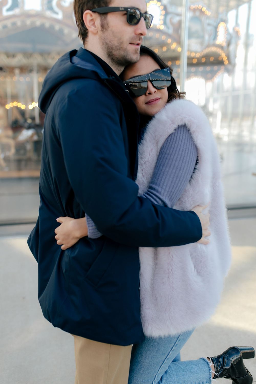 Couple embracing outdoors near a carousel, both wearing sunglasses and winter jackets on a romantic date.