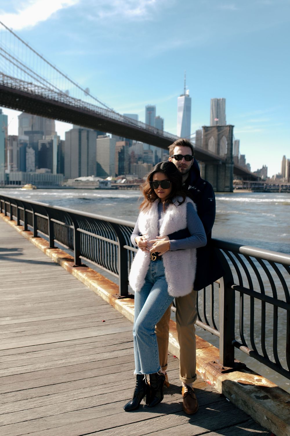 Couple posing on Brooklyn Bridge Park waterfront boardwalk with NYC skyline and Brooklyn Bridge in background.