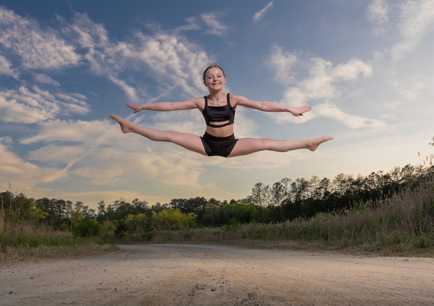 Young dancer performs a perfect split leap mid-air outdoors against a dramatic blue sky with clouds.