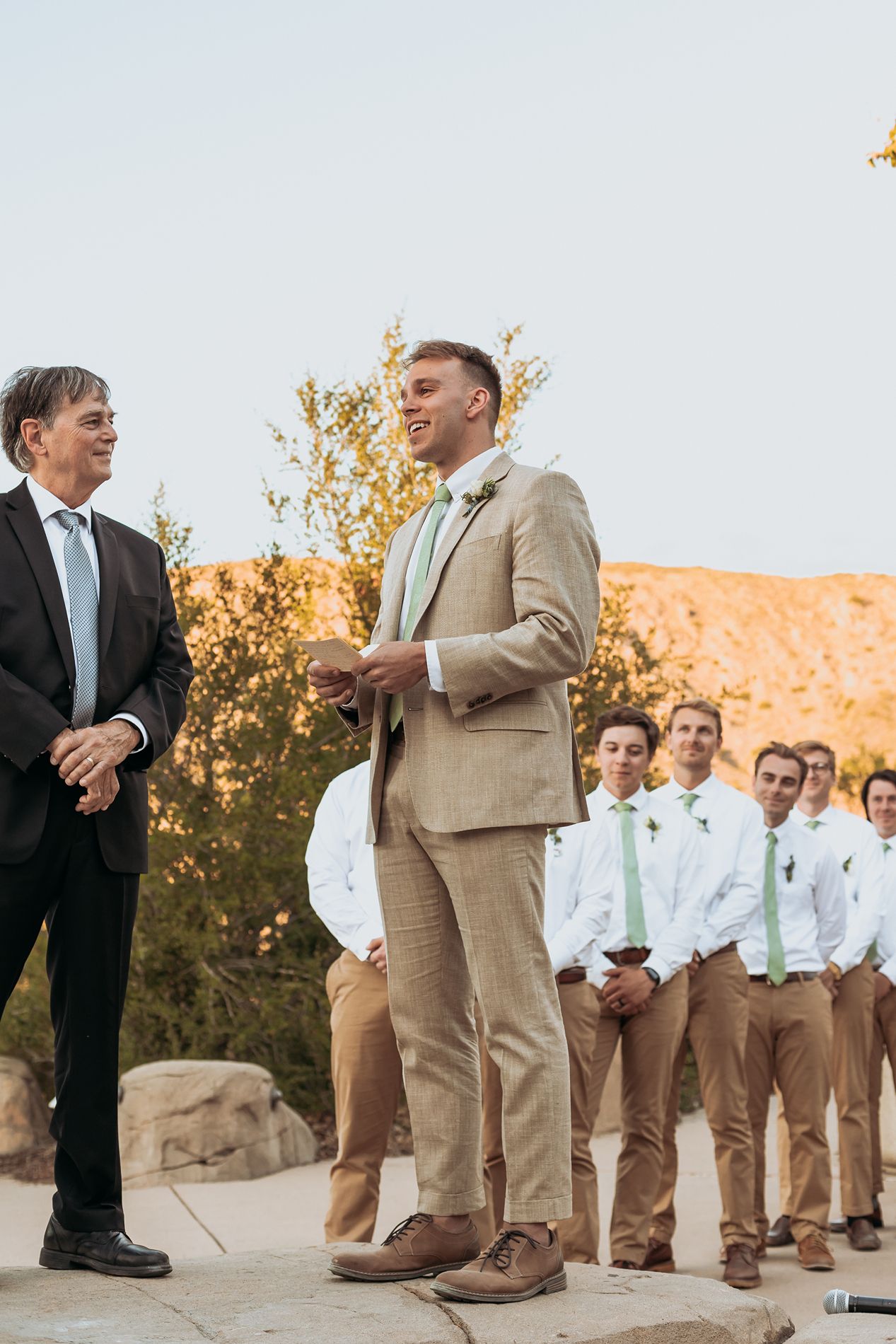 Groom in tan suit with mint tie laughing at outdoor desert wedding ceremony with groomsmen in white shirts at Mission Trails.