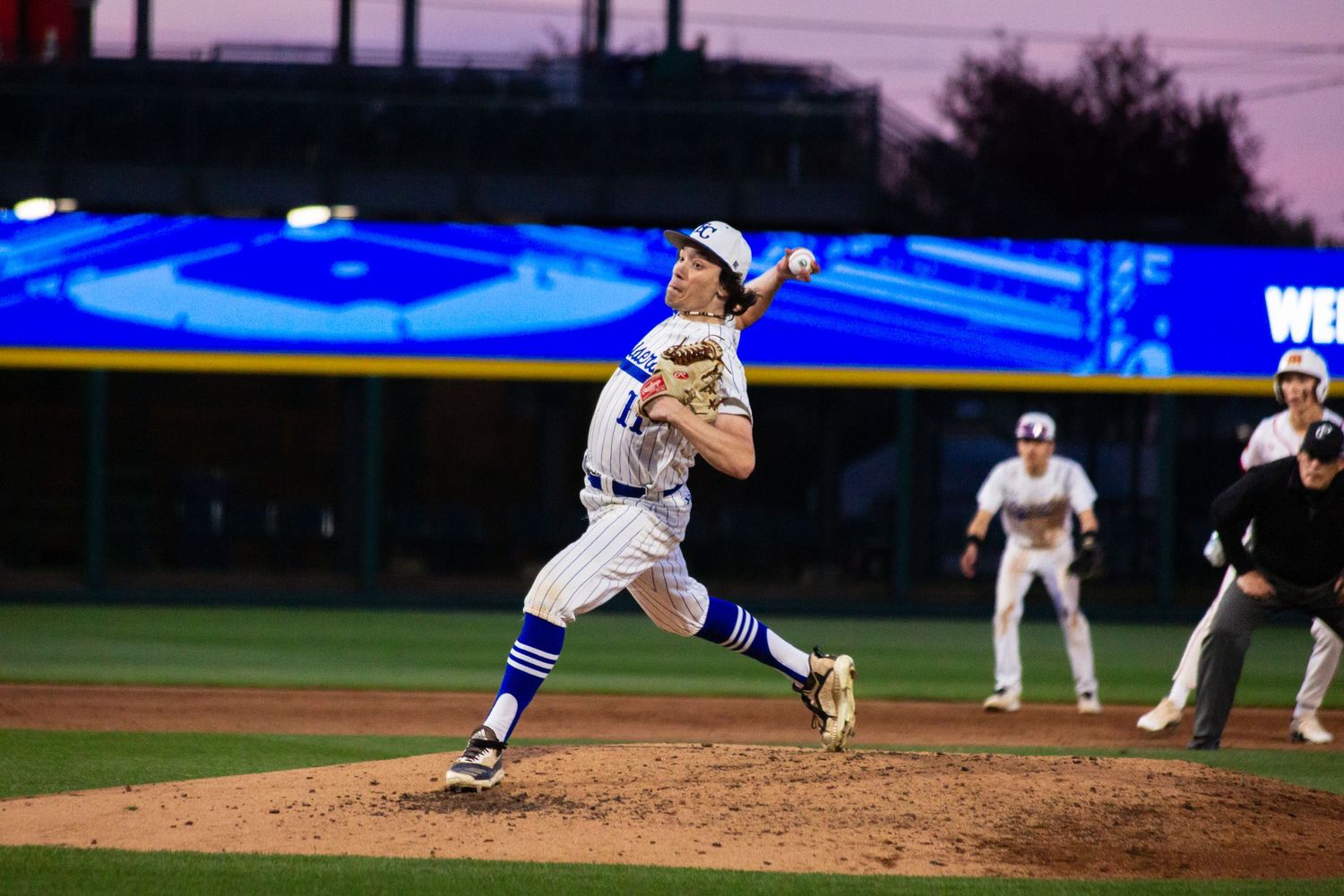Baseball pitcher in blue and gold uniform mid-throw during a night game under stadium lights.